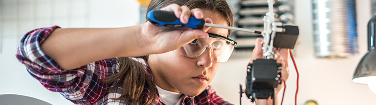 Image of a grade-school aged boy in a classroom with safety goggles on, using a screwdriver to work on a small mechanical device.