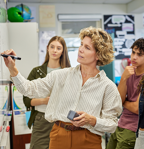 Image shows a teacher writing on the whiteboard as a teen girl watches.