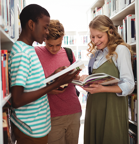 three students in a library standing between bookshelves with books in hand