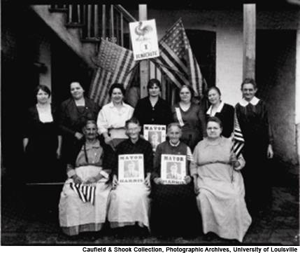 Black and white photo of women suffragists seated together, holding political signs and American flags.